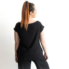 A woman with long, straight, reddish-brown hair in a high ponytail stands with her back to the camera, wearing the Curve Hem T-Shirt by KOBOMO Bamboo in black and black pants against a plain white background.
