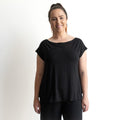 A woman with light skin and brown hair in a ponytail smiles at the camera, wearing the KOBOMO Bamboo Curve Hem T-Shirt and black pants, standing against a plain white background.