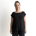 A woman with light skin and brown hair in a ponytail smiles at the camera, wearing the KOBOMO Bamboo Curve Hem T-Shirt and black pants, standing against a plain white background.