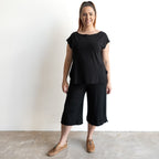 A woman with brown hair in a ponytail smiles against a white wall, wearing the Curve Hem T-Shirt by KOBOMO Bamboo paired with black wide-leg cropped pants and brown lace-up shoes.