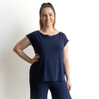 A woman with long brown hair in a ponytail smiles at the camera, wearing the Curve Hem T-Shirt by KOBOMO Bamboo with matching pants. She stands against a plain white background, one hand resting on her hip.