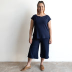 A woman smiles against a white wall, wearing the KOBOMO Bamboo Curve Hem T-Shirt in navy with matching wide-leg cropped pants and brown lace-up shoes.