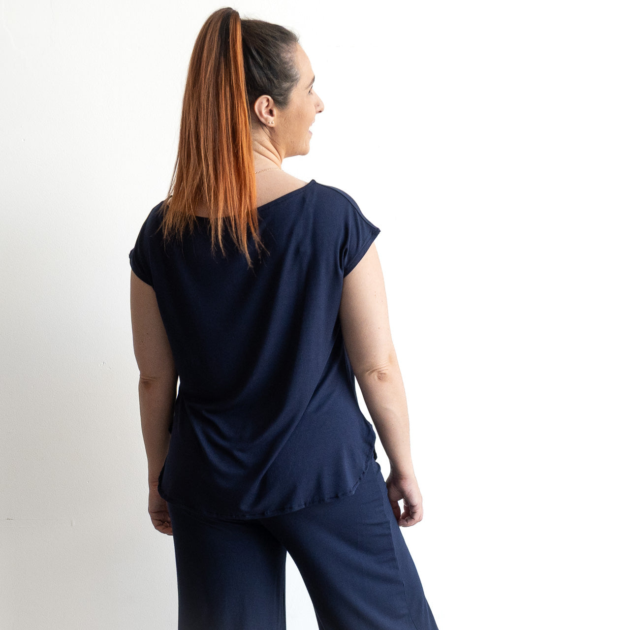 A woman with long brown hair in a ponytail stands with her back to the camera, wearing the navy Curve Hem T-Shirt by KOBOMO Bamboo and matching pants against a plain white background.