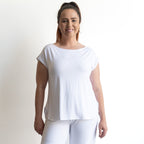 A woman with long brown hair in a high ponytail is smiling against a plain white background, wearing the Curve Hem T-Shirt by KOBOMO Bamboo with short sleeves and paired with white pants.