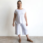 A smiling woman with brown hair tied back stands against a white wall, wearing the Curve Hem T-Shirt by KOBOMO Bamboo—a loose-fitting, short-sleeve white top—paired with white wide-leg cropped pants and brown lace-up shoes.