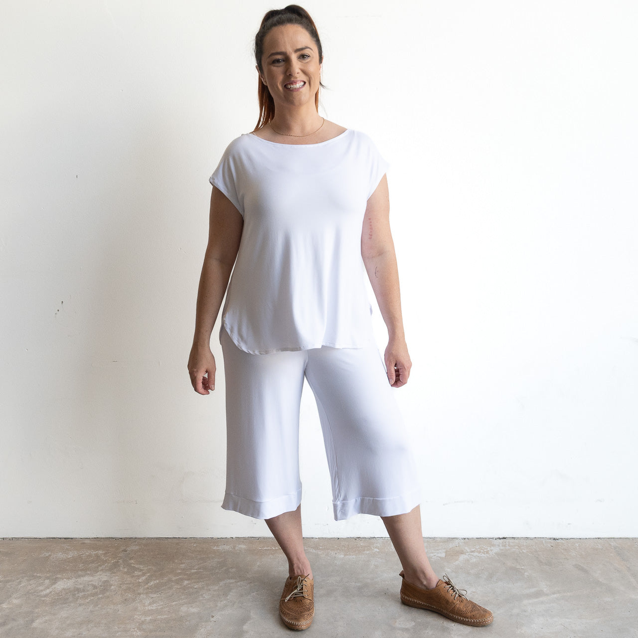 A smiling woman with brown hair tied back stands against a white wall, wearing the Curve Hem T-Shirt by KOBOMO Bamboo—a loose-fitting, short-sleeve white top—paired with white wide-leg cropped pants and brown lace-up shoes.
