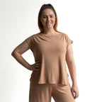 A woman with long brown hair in a high ponytail smiles against a plain white background. She wears the Curve Hem T-Shirt by KOBOMO Bamboo—a loose, breathable women's top with short sleeves—paired with matching pants, one hand on her hip.