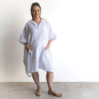 The Noosa Beach Hoodie in white cotton, knee-length with elbow sleeves, front pockets, and a relaxed fit. Tan slip-on shoes.